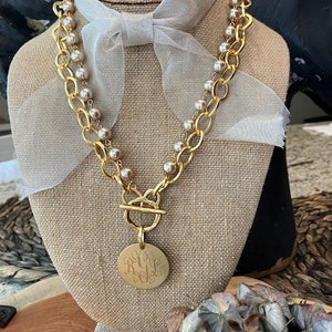 May include: A gold chain necklace with a large round pendant featuring a monogram. The necklace is layered with a strand of faux pearls.