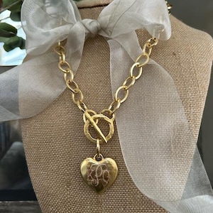 May include: A gold chain necklace with a large, heart-shaped pendant. The pendant is engraved with the initials "K.P.O.". The necklace is displayed on a mannequin with a white ribbon.
