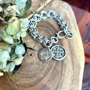 May include: A silver-toned chain link bracelet featuring two circular charms. One charm is a flat disc engraved with the letters MGM in a script font, and the other charm displays a raised, textured cross design. The bracelet has a lobster clasp.