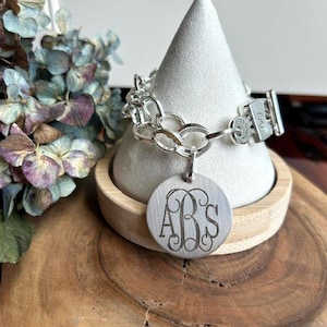 May include: A silver-toned chain link bracelet featuring a round, brushed metal pendant engraved with the monogram letters ABS in a decorative script font. The bracelet includes a rectangular toggle clasp closure with a small circular charm.