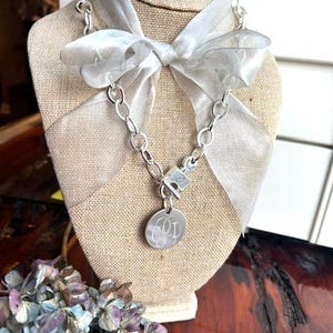 May include: A silver-toned chain necklace featuring a toggle clasp and a round pendant engraved with the letters GPD. The necklace is accented with a translucent, shimmering silver ribbon tied into a bow at the center of the chain.