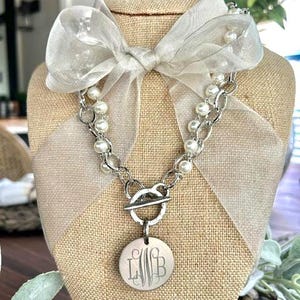 May include: A silver-toned chain necklace featuring a toggle clasp, white faux pearls, and a sheer white organza ribbon tied in a bow. A circular silver-toned pendant hangs from the toggle, engraved with the monogram letters LWB in a script font.