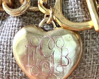 May include: A gold-toned metal heart-shaped pendant attached to a thick, textured chain link necklace. The center of the heart features an engraved monogram with the letters E, C, and B in a decorative, swirling script font style.