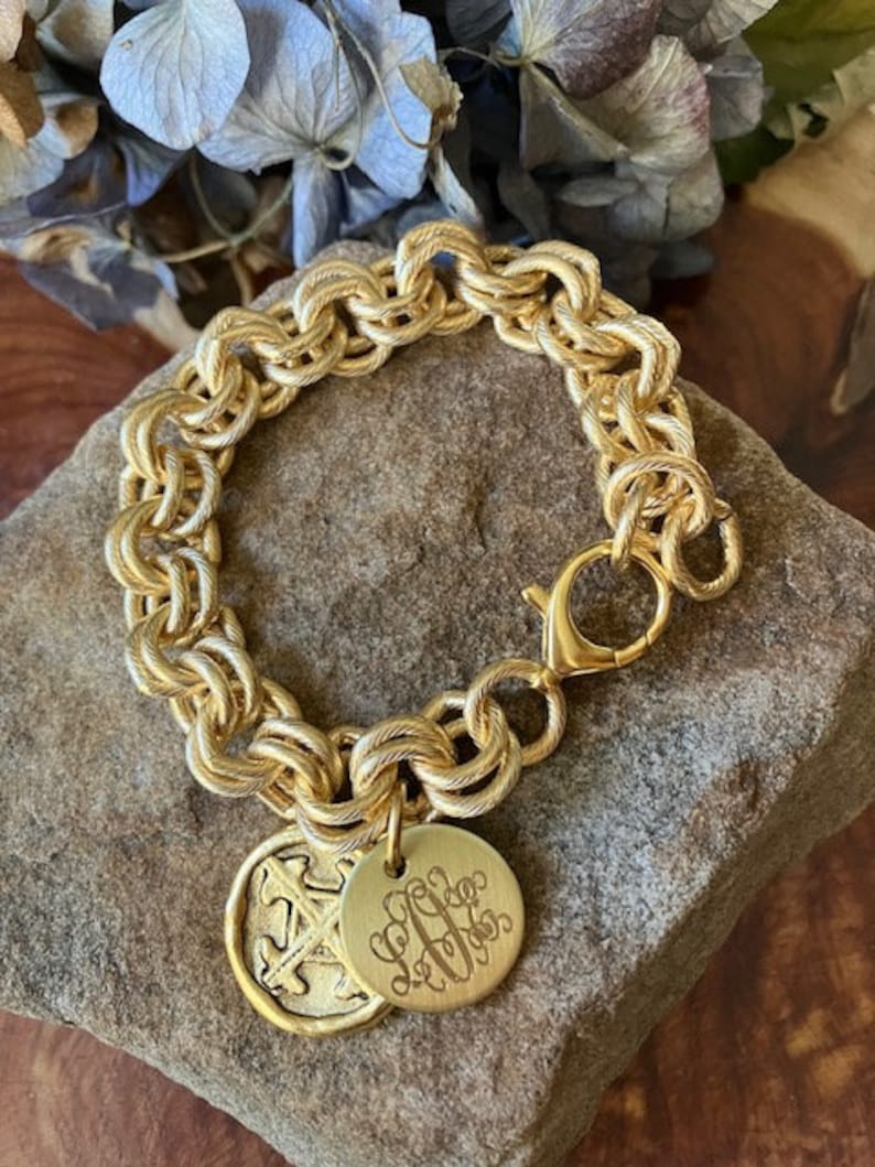 May include: A gold chain bracelet with a lobster clasp and two charms. One charm is a round disc with a monogram, and the other charm is a round disc with a cross design.