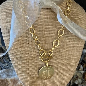 May include: A gold chain necklace with a large, round pendant featuring the monogram "WOS". The chain is thick and chunky, and the pendant is attached to a toggle clasp.