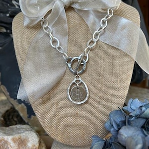 May include: A silver chain necklace with a toggle clasp and a round pendant with the initials "CHX" engraved on it. The necklace is displayed on a mannequin with a light brown fabric base and a white sheer ribbon bow.