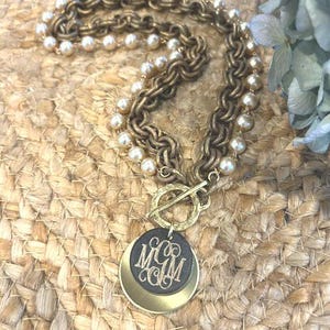May include: A necklace featuring a double-strand chain of antique gold-toned metal links and cream-colored pearls. The piece includes a toggle clasp and a layered pendant consisting of a black circular disc with the monogram MGM and a gold disc.