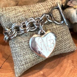 May include: A silver-toned chain link bracelet featuring a toggle clasp and a large, smooth heart-shaped pendant. The pendant is engraved with the ornate, cursive monogram letters NMB. The metal has a polished finish with visible textured links.