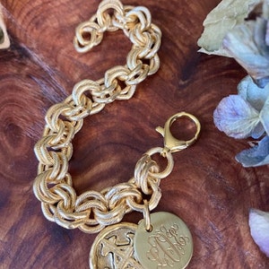 May include: A gold chain bracelet with a gold charm featuring a monogram and a gold charm with a textured design.