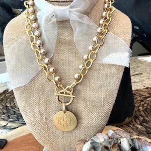 May include: A layered necklace featuring a gold-toned chain and a strand of champagne-colored pearls. The necklace includes a gold-toned toggle clasp and a circular gold-toned pendant engraved with the monogram BGL, accented by a sheer ribbon.