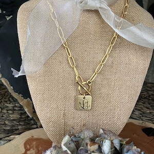 May include: A gold chain necklace with a square pendant featuring the monogram "AJL". The necklace is displayed on a fabric-covered mannequin with a white ribbon.