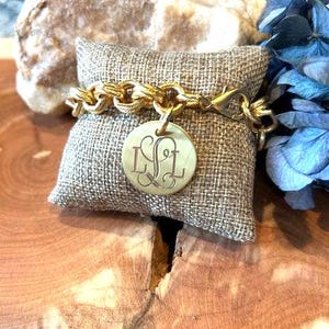 May include: A gold-toned chain link bracelet featuring a round pendant engraved with the letters LSL in a decorative script font. The bracelet includes a lobster claw clasp and is displayed on a small, square, textured beige fabric pillow.