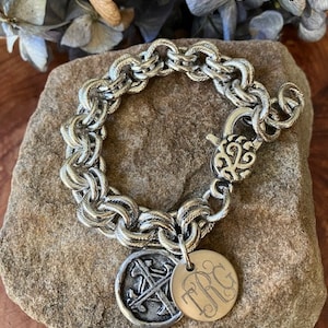 May include: A silver-toned bracelet with a double-strand chain design. The bracelet features a decorative clasp and two round charms. One charm has an anchor design, and the other has the initials "TRG". The bracelet is displayed on a stone surface.