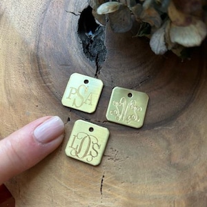 May include: Three gold-colored metal square tags with engraved initials. The tags are on a wooden surface.
