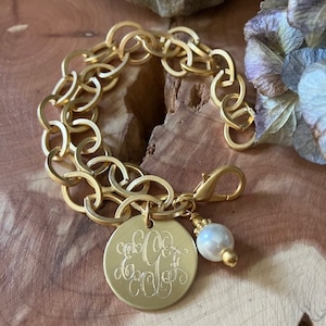 May include: A gold chain bracelet with a round charm featuring a monogram and a pearl accent.