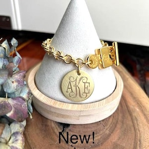 May include: A gold-toned chain bracelet featuring a round, brushed-finish pendant engraved with the monogram letters AKE in a decorative script font. The bracelet includes a rectangular toggle clasp closure and is displayed on a cone-shaped stand.