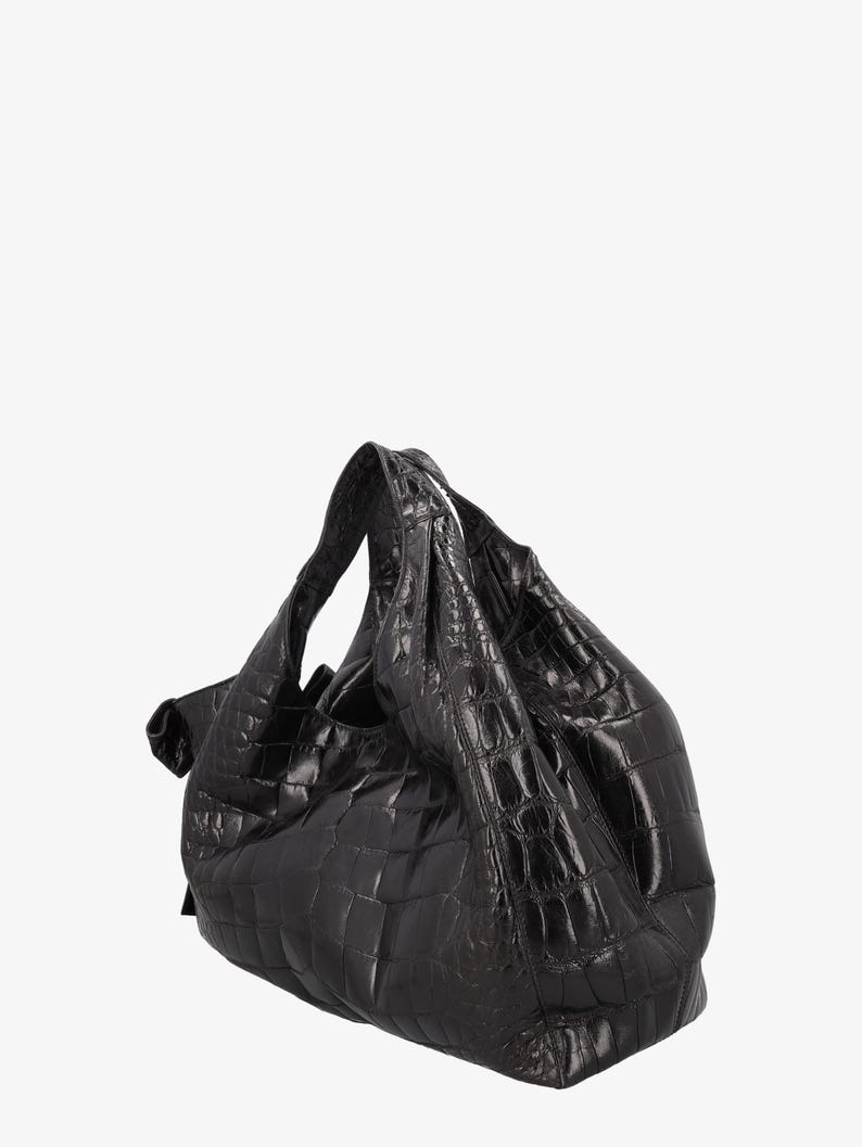 May include: A black, textured handbag with a crocodile-like pattern. The bag has a soft, slouchy shape with a single handle. The material appears to be leather or a leather-like material. The bag is set against a white background.