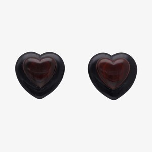 May include: Two heart-shaped earrings. Each earring features a dark red heart set within a black heart-shaped frame. The earrings are displayed against a white background.