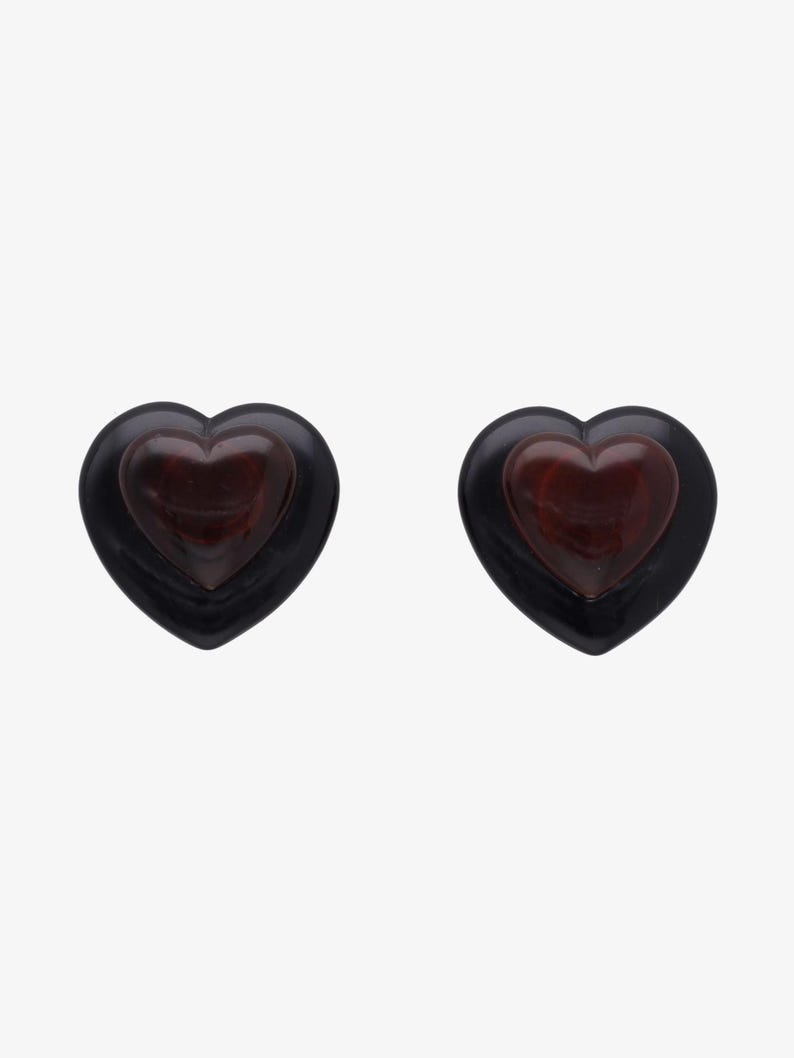 May include: Two heart-shaped earrings. Each earring features a dark red heart set within a black heart-shaped frame. The earrings are displayed against a white background.