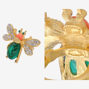 Op de afbeelding: Gouden servetring in de vorm van een bij met groene en koraalkleurige accenten. De ring is gemarkeerd met "Roberta Diane" aan de onderkant.