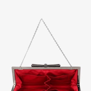Pode incluir: Embreagem prateada aberta com interior de cetim vermelho. A embreagem apresenta um la&ccedil;o decorativo com enfeites pretos e prateados e uma al&ccedil;a de corrente prateada. O interior tem v&aacute;rios bolsos.