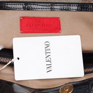 May include: Close-up of a black leather handbag interior with a gold zipper and a red Valentino label. A white tag with "VALENTINO" printed vertically is attached. The bag's interior is tan.