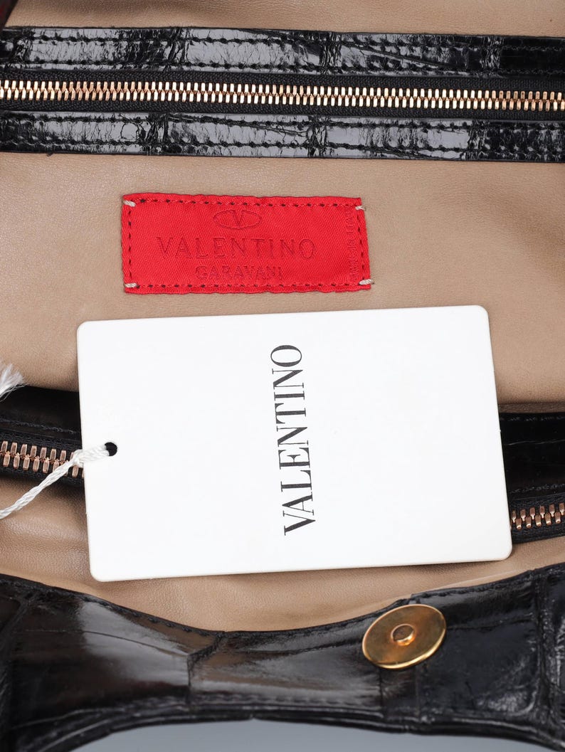 May include: Close-up of a black leather handbag interior with a gold zipper and a red Valentino label. A white tag with "VALENTINO" printed vertically is attached. The bag's interior is tan.