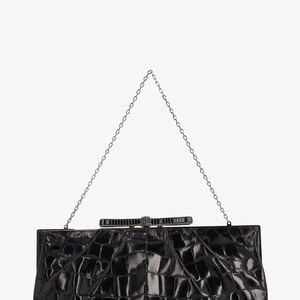 Pode incluir: Uma clutch retangular preta com um padr&atilde;o texturizado semelhante ao de crocodilo. Possui uma corrente prateada e um fecho decorativo com design de la&ccedil;o. Um acess&oacute;rio formal.