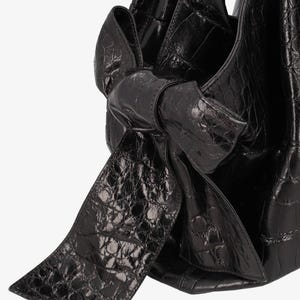 May include: Black leather handbag with a bow detail. The bag has a textured, crocodile-like pattern. The bow is large and prominent, adding a decorative element to the bag's design. The bag appears to be made of a shiny, high-quality leather.