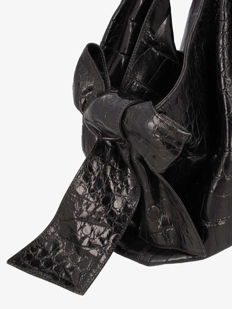 May include: Black leather handbag with a bow detail. The bag has a textured, crocodile-like pattern. The bow is large and prominent, adding a decorative element to the bag's design. The bag appears to be made of a shiny, high-quality leather.