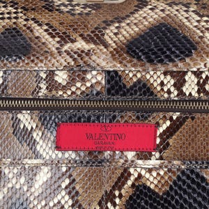 Op de afbeelding: Close-up van een luxe handtas met een slangenleerpatroon in bruin, zwart en cr&egrave;me. De tas heeft een zilverkleurige sluiting en een rood label met het "Valentino Garavani" logo.