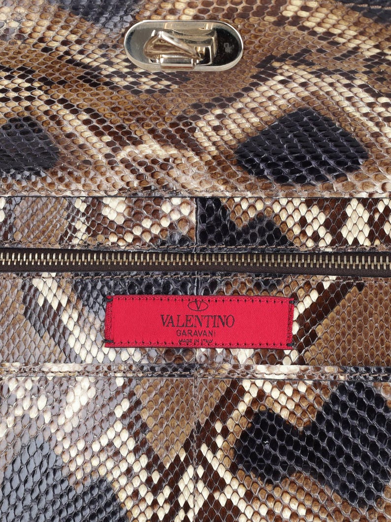 Op de afbeelding: Close-up van een luxe handtas met een slangenleerpatroon in bruin, zwart en cr&egrave;me. De tas heeft een zilverkleurige sluiting en een rood label met het "Valentino Garavani" logo.