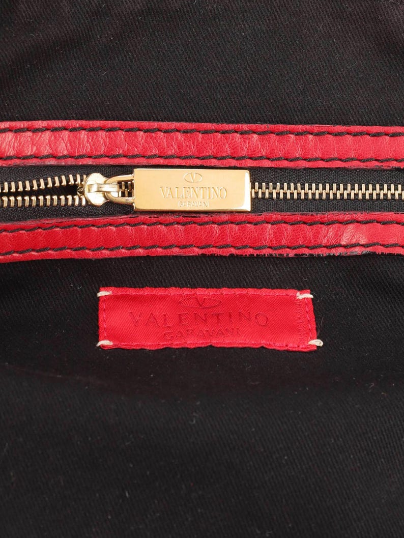 Op de afbeelding: Close-up van zwarte stof met rode leren afwerking en een gouden rits. Een rechthoekige gouden plaat met de merknaam "VALENTINO GARAVANI" staat boven de rits. Een rood label met de merknaam is op de stof gestikt.