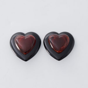May include: Two heart-shaped earrings. Each earring has a black outer heart and a smaller, reddish-brown heart in the center. The earrings are set against a white background, highlighting their glossy finish and bold design.