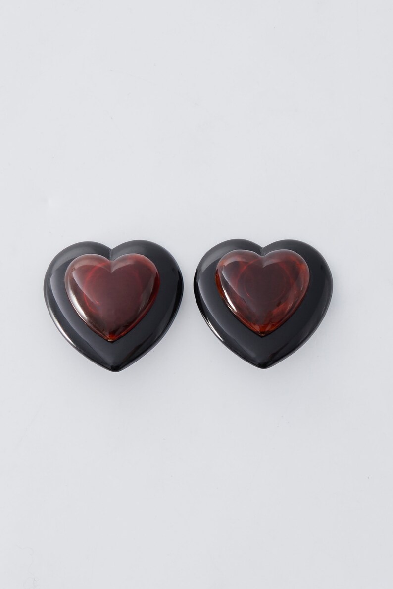 May include: Two heart-shaped earrings. Each earring has a black outer heart and a smaller, reddish-brown heart in the center. The earrings are set against a white background, highlighting their glossy finish and bold design.