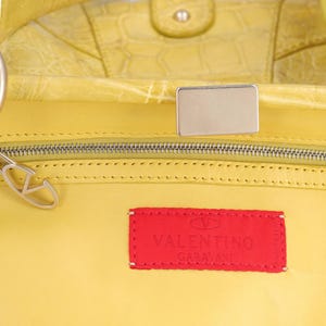 以下が含まれることがあります： シルバーのジッパーと金具が付いた、黄色の革製ハンドバッグのクローズアップ。 「Valentino Garavani」のロゴが入った赤い長方形のタグがバッグに縫い付けられています。 バッグはクロコダイルのような質感のパターンです。