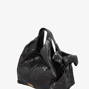 May include: A black leather handbag with a crocodile pattern. The bag has a large, slouchy shape with a top handle and a decorative bow on the side. A small, rectangular gold-colored metal plate is on the front.