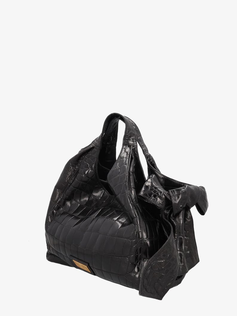 May include: A black leather handbag with a crocodile pattern. The bag has a large, slouchy shape with a top handle and a decorative bow on the side. A small, rectangular gold-colored metal plate is on the front.