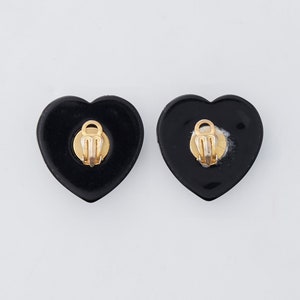 May include: A pair of black heart-shaped clip-on earrings. The earrings have a glossy finish and gold-colored clips on the back. The earrings are displayed on a white background.