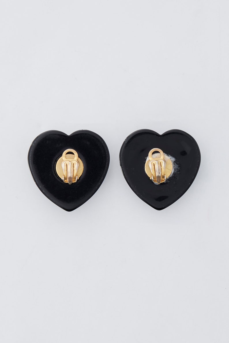 May include: A pair of black heart-shaped clip-on earrings. The earrings have a glossy finish and gold-colored clips on the back. The earrings are displayed on a white background.