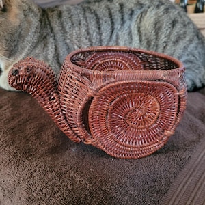 May include: A dark brown woven basket shaped like a snail. The basket features a coiled shell design on the side and a protruding head section with a small dark eye detail. The structure is crafted from thin, flexible reeds woven into a pattern.