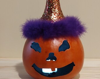 May include: A carved orange gourd lantern with a jack-o'-lantern face featuring cutouts for eyes, a nose, and a smiling mouth. The gourd wears a conical hat covered in gold glitter with a fluffy purple feather trim around the base of the hat.