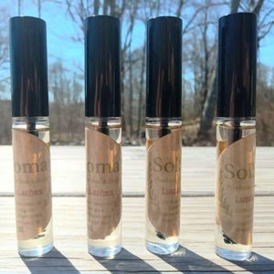 May include: Four clear glass roller bottles with black caps. Each bottle has a tan label with the words "Aroma Solutions" and "Lashes" printed on it. The bottles are filled with a clear liquid.