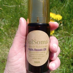 May include: A brown glass bottle with a gold cap, labeled "SolSoma Night Repair Oil." The label includes text about the product's ingredients and volume, 50ml. The bottle is held against a backdrop of green grass and yellow flowers.