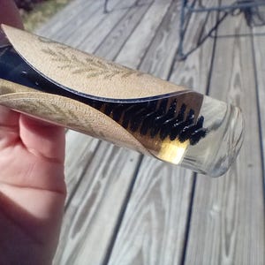 May include: A clear glass bottle containing a brush with black bristles and a light-colored liquid. The bottle is partially wrapped with a tan paper label with a leaf design.