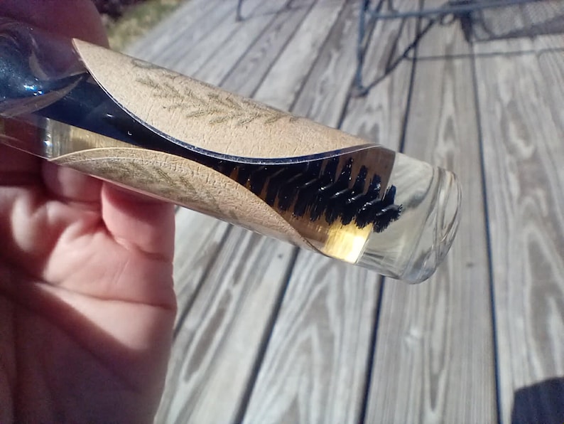 May include: A clear glass bottle containing a brush with black bristles and a light-colored liquid. The bottle is partially wrapped with a tan paper label with a leaf design.