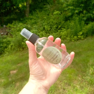 May include: A clear glass spray bottle with a black and white cap, held in a hand. The bottle has a label that reads "SolSoma Cooling + Calming Summerspray 4 oz". The background is blurred green foliage.