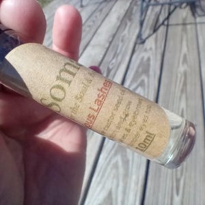 May include: A clear glass roller bottle with a black cap, labeled "Som" and "Lashes." The label is tan with black text, including "castor seed oil" and "10ml." The bottle is held against a wooden surface.