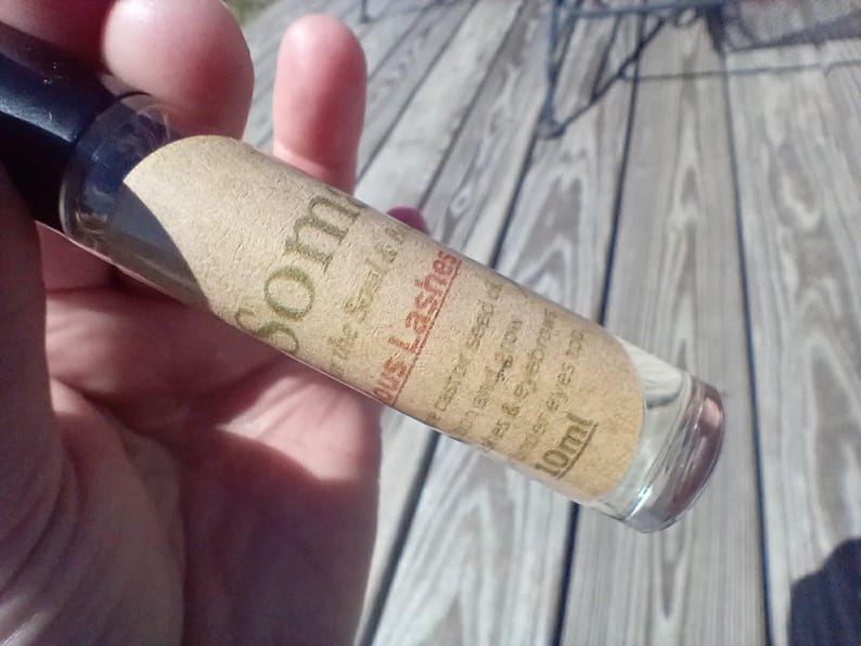May include: A clear glass roller bottle with a black cap, labeled "Som" and "Lashes." The label is tan with black text, including "castor seed oil" and "10ml." The bottle is held against a wooden surface.