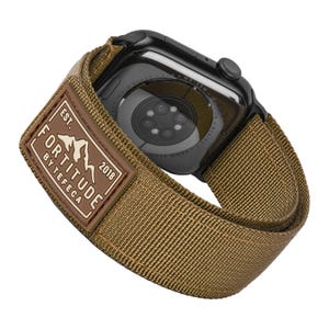 Puede incluir: Una correa de reloj de nailon marrón con un Apple Watch negro. La correa tiene un parche de cuero marrón con el texto "FORTITUDE BY TEFECA EST. 2018".